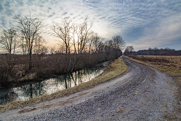Dirt and gravel road curving around near a stream with a blue, cloudy sky